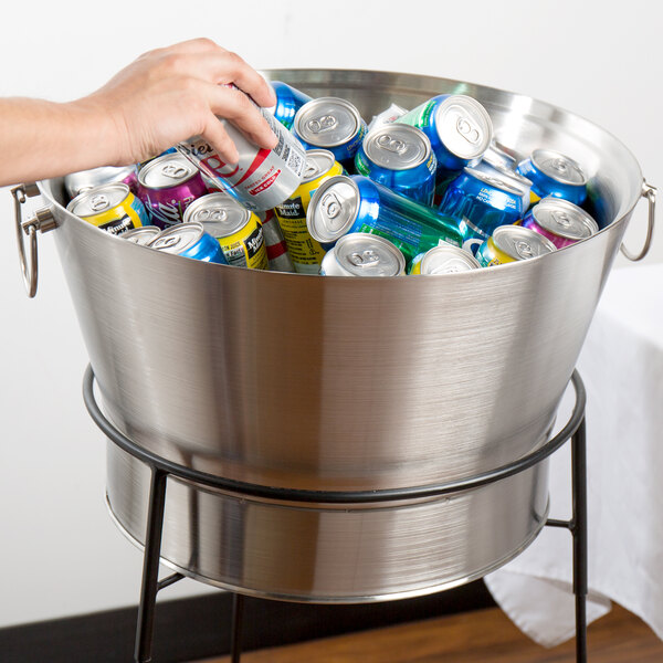 A hand reaching for a blue and white soda can in a Tablecraft stainless steel beverage tub.