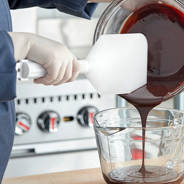 A gloved hand uses a white Carlisle Sparta paddle to pour chocolate into a bowl.