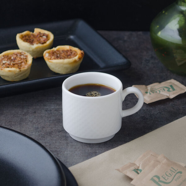 A white Villeroy & Boch porcelain stackable cup filled with brown liquid sits on a table with small pastries.