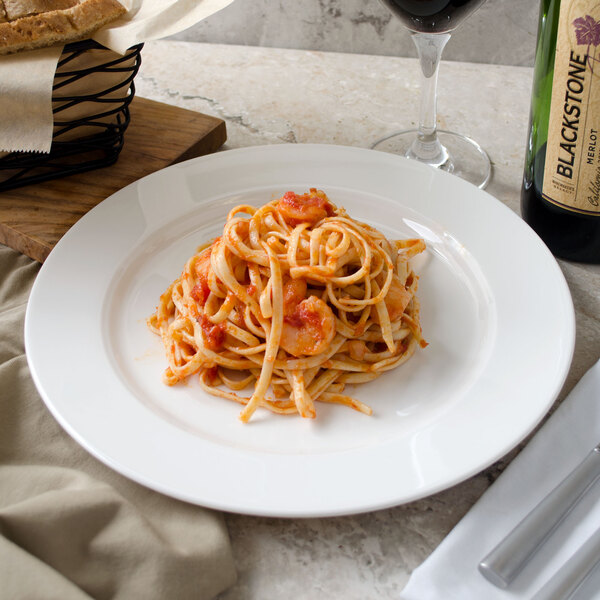 A white porcelain plate of pasta with shrimp and a glass of wine on a table.