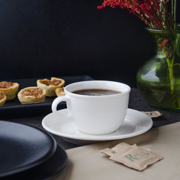 A Villeroy & Boch white porcelain saucer with a cup of coffee on it on a table next to a vase of red flowers.