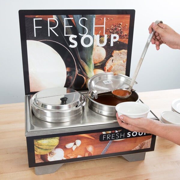 A person using a ladle to pour soup from a Vollrath Tuscan soup merchandiser into a bowl.