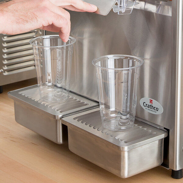 A person using a Crathco refrigerated beverage dispenser to fill two plastic cups.