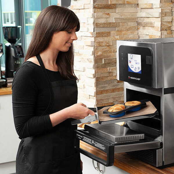 A woman wearing a black apron putting food into a Merrychef countertop oven on a kitchen counter.