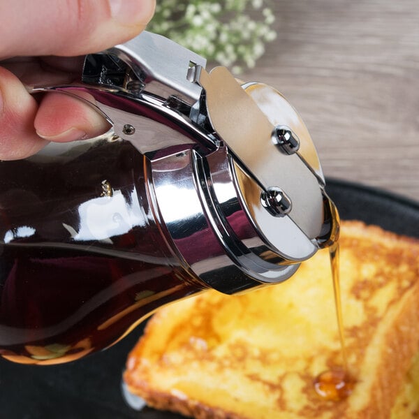 A person pouring syrup onto a piece of french toast using a Tablecraft glass syrup dispenser with a chrome-plated top.
