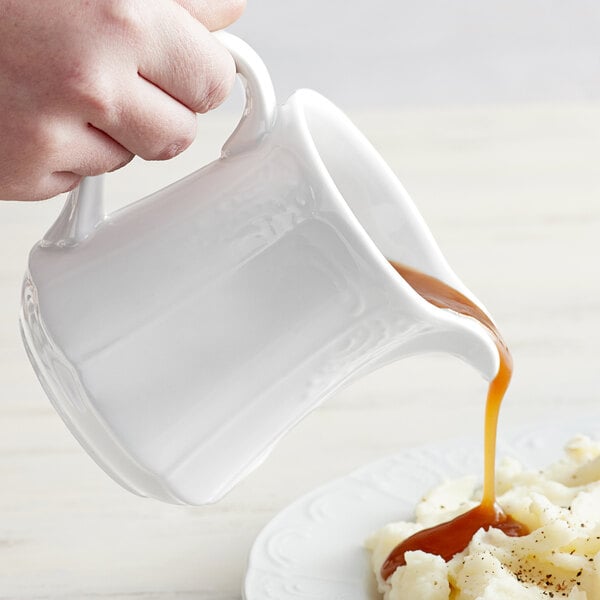 A person pouring brown gravy from a Tuxton Bright White China gravy boat into a plate of food.