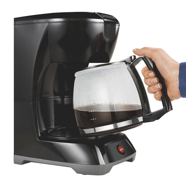 A hand holding a black Hamilton Beach coffee maker pouring coffee into a glass pot.