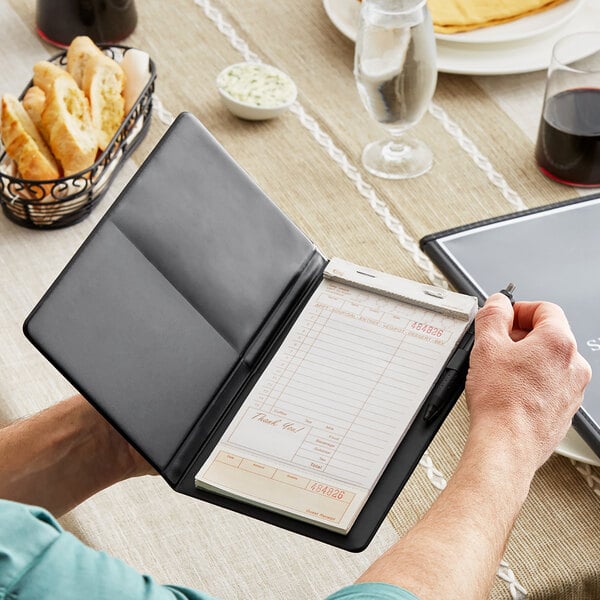 A person holding a H. Risch, Inc. black vinyl server book and a black pen with a receipt inside.