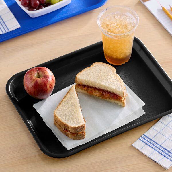 A black plastic fast food tray holding a peanut butter and jelly sandwich, an apple, and a cup of iced beverage.