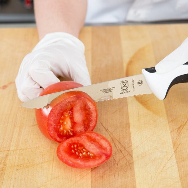 A person in gloves uses a Mercer Culinary Millennia utility knife to cut a tomato on a counter.