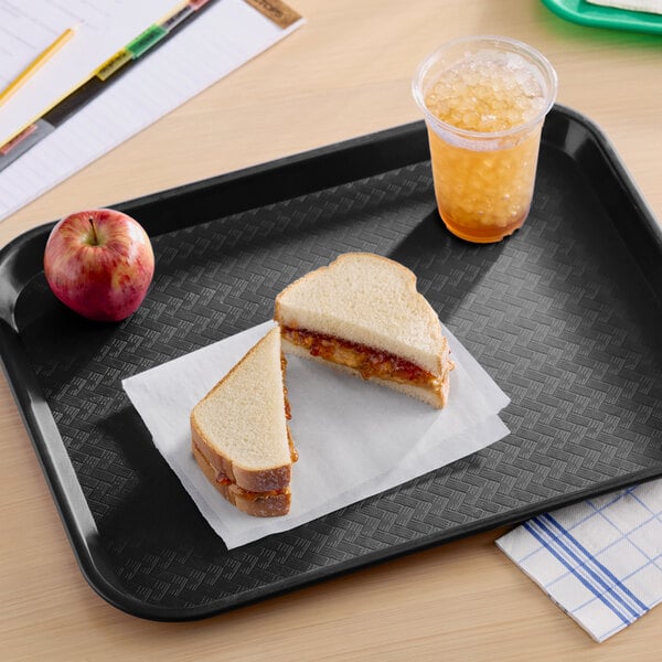 A black plastic fast food tray holding a peanut butter and jelly sandwich, an apple, and a cup of iced drink.