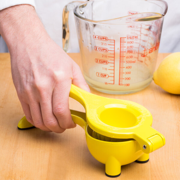 A hand using a Tablecraft yellow zinc alloy lemon squeezer to juice a lemon into a measuring cup.