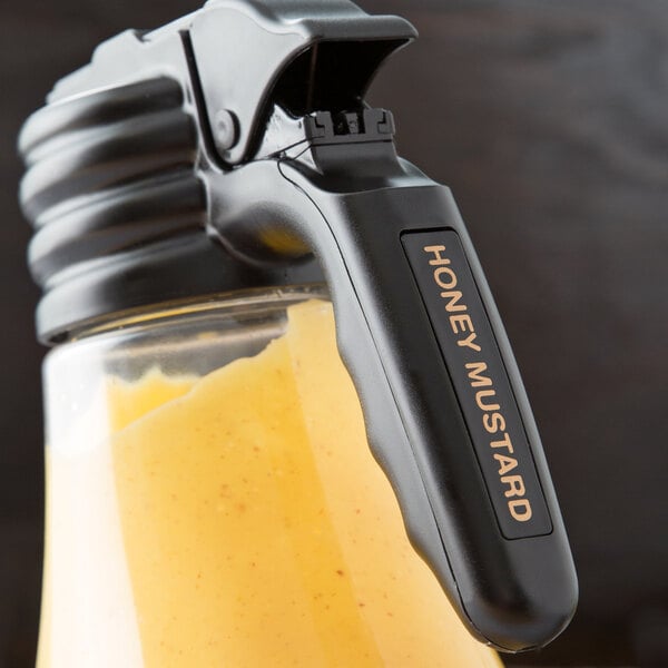 A Tablecraft black polypropylene print dispenser tag on a container of honey mustard on a table.