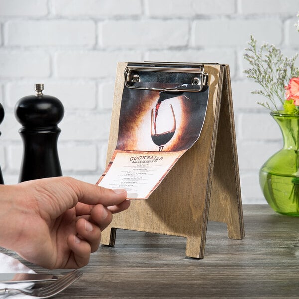 A hand holding a Menu Solutions weathered walnut wood sandwich menu board tent with a clip on a wooden table.