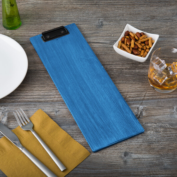 A blue wooden menu clip board measuring 4 1/4 by 14 inches, placed on a table setting.