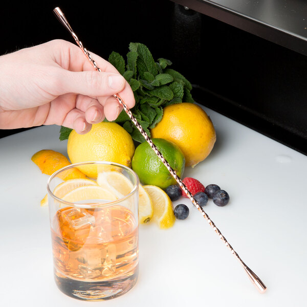 A hand using a Barfly copper plated double end stirrer to mix a drink with lemon slices.