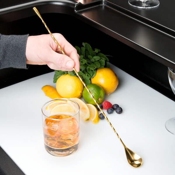 A hand holding a Barfly gold plated bar spoon over a glass of iced lemonade.