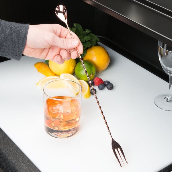 A hand using a Barfly copper plated bar spoon with a fork end to stir a glass of liquid with ice and a slice of lemon.