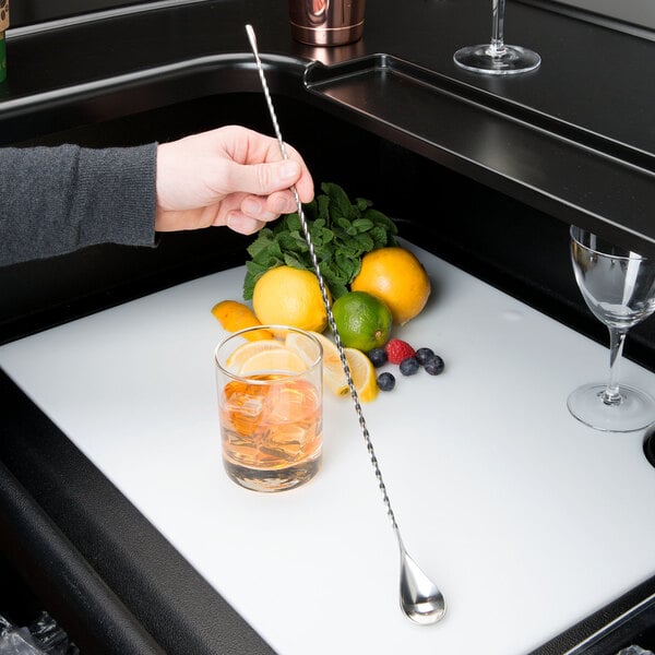 A person using a Barfly stainless steel classic bar spoon to stir a cocktail with ice and a lemon.