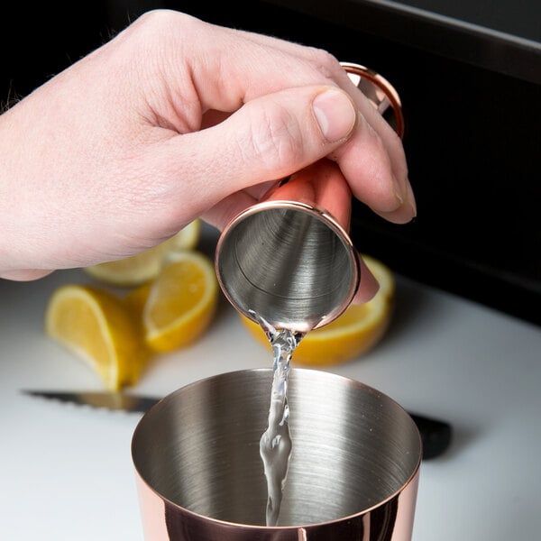 A person using a Barfly copper-plated Japanese style jigger to pour liquid into a metal cup.