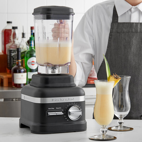 A person in a black apron using a KitchenAid Matte Black blender on a counter.