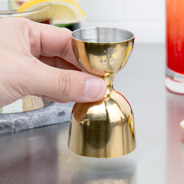 A hand using a Barfly gold-plated metal jigger to pour yellow liquid.
