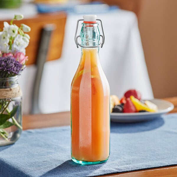 An Arcoroc Swing Top Tinted Green Glass Bottle filled with orange juice on a table.