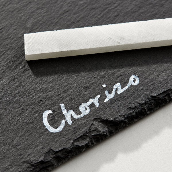 A black slate tray with the word 'Chorizo' written on it in white chalk and a piece of soapstone chalk resting on the tray.