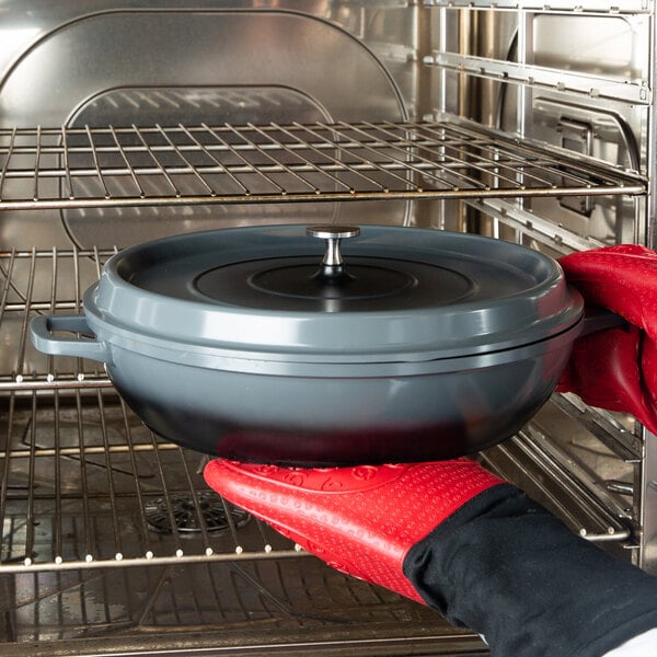 A person in red gloves holding a GET Heiss gray enamel coated cast aluminum brazier with a lid in an oven.