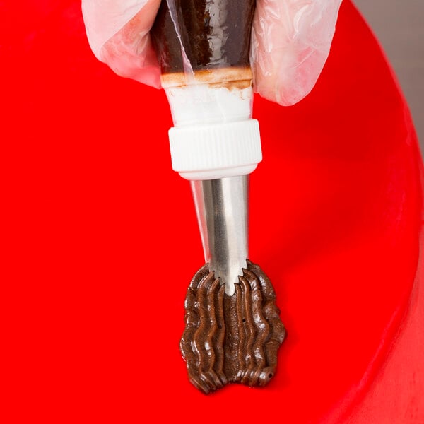 A hand holding an Ateco pastry bag with brown frosting over a red surface.