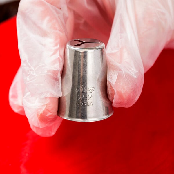 A hand in a glove holding a silver metal Ateco heart piping tip.
