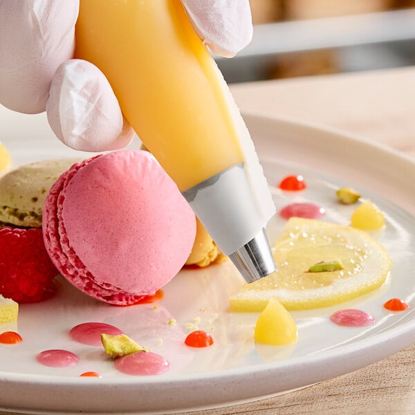 A hand using an Ateco plain piping tip to pipe cream onto a pink macaroon.