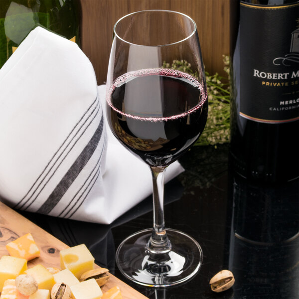An Arcoroc wine glass filled with red wine on a table with a bottle of cheese and nuts.