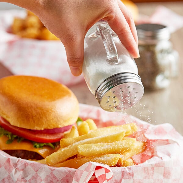 A hand using an Acopa mini mason jar salt shaker to sprinkle salt on a burger and fries.