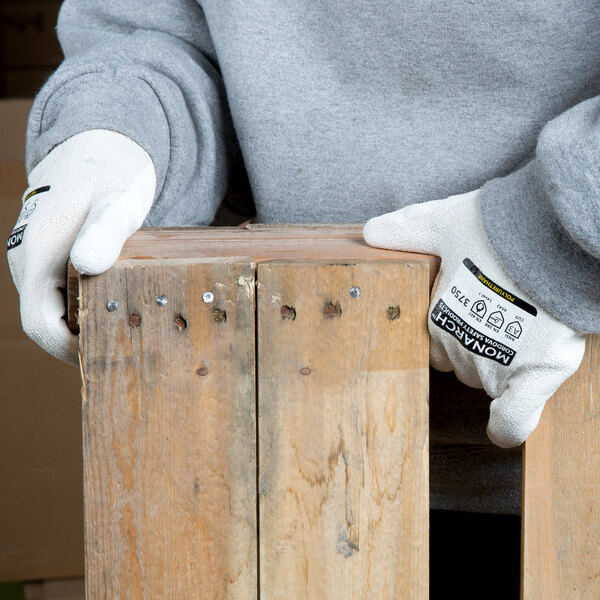 A pair of white cut resistant gloves with a white polyurethane palm coating being worn by a person handling wooden boards.