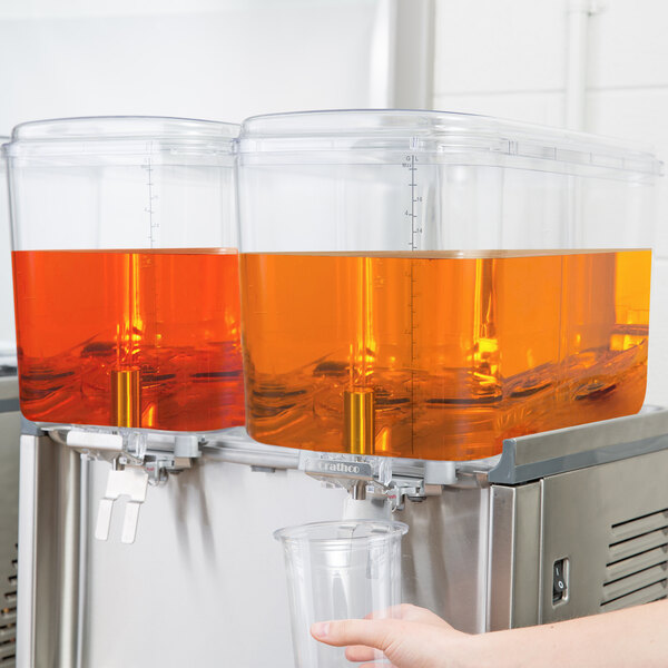 A hand pouring orange juice from a Crathco beverage dispenser into a plastic cup.