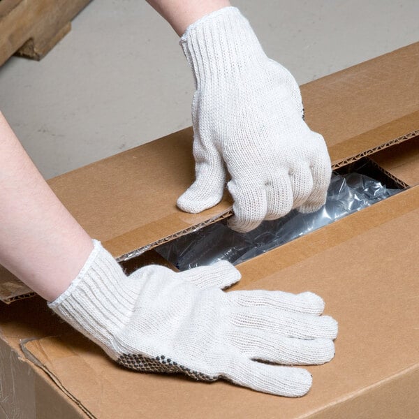 A person wearing Cordova economy work gloves with black PVC dots opening a cardboard box.