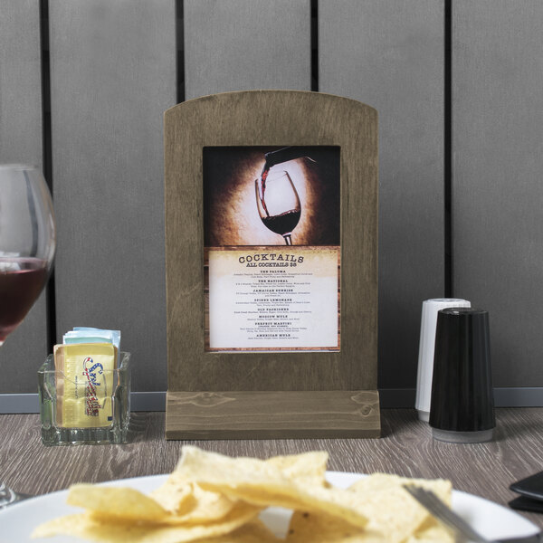 A Menu Solutions weathered walnut wood menu tent on a table with a plate of chips and a wine glass.