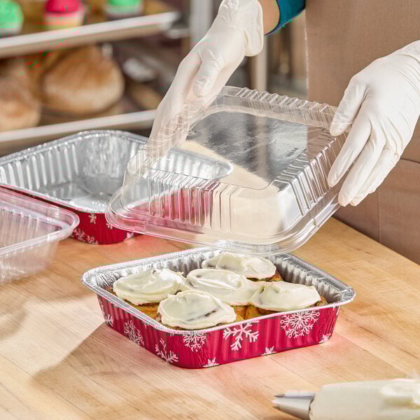 An 8-inch square holiday-themed foil cake pan with a clear dome lid, containing frosted baked goods.