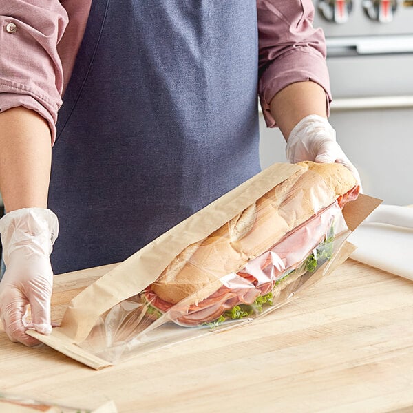 A person in a blue apron holding a sandwich in a Bagcraft Dubl View Kraft paper bag with a window.