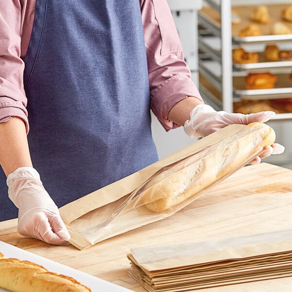 A woman in a blue apron holding a Bagcraft Packaging Dubl View ToGo! Kraft window bag with a baguette inside.