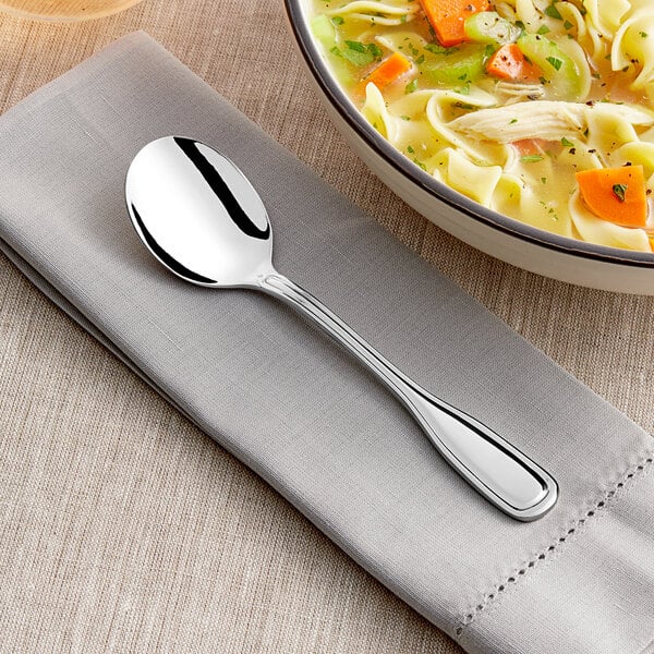 An Acopa Scottdale stainless steel bouillon spoon on a napkin next to a bowl of soup.