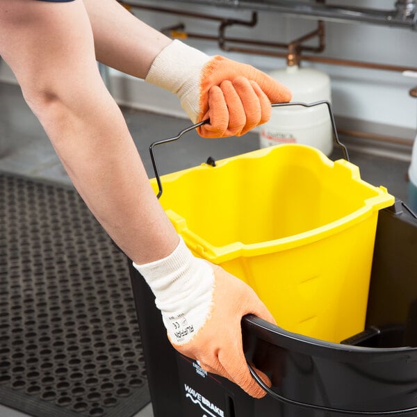 A person wearing orange Cordova Ruffian gloves holding a yellow bucket.