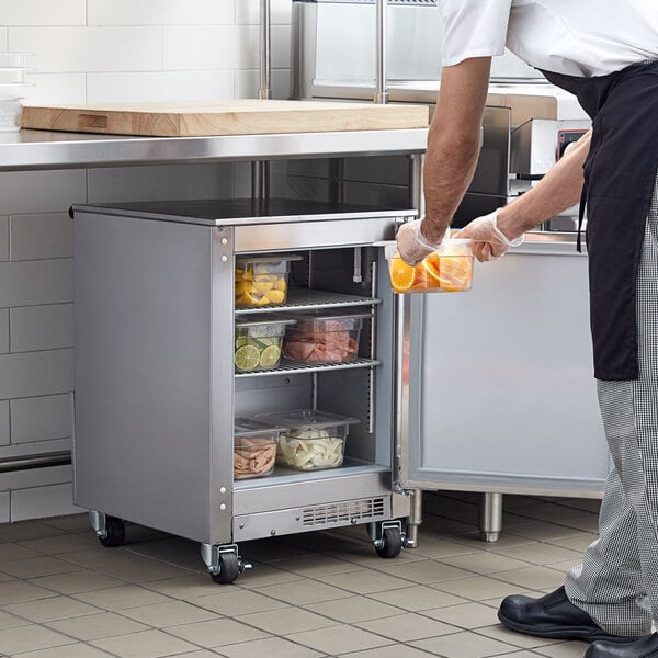 A man in a white shirt putting a container of oranges into a Beverage-Air undercounter refrigerator in a school kitchen.