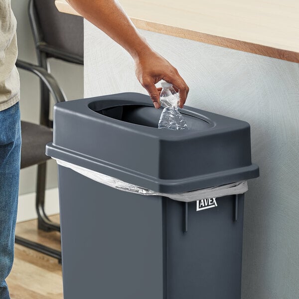 A hand putting a plastic bottle into a Lavex Gray Slim Rectangular Trash Can.