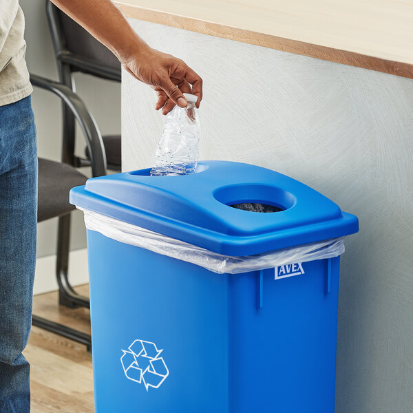 A person putting a plastic bottle into a blue Lavex Slim rectangular recycling trash can with a plastic bag over it.