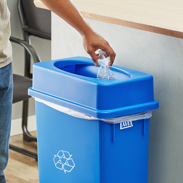 A person putting a plastic bottle into a blue recycling bin with a Lavex Blue Slim Rectangular Trash Can Drop Shot Lid.