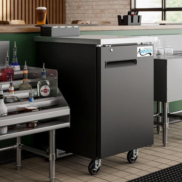 An Avantco black back bar refrigerator on a counter filled with bottles of alcohol.