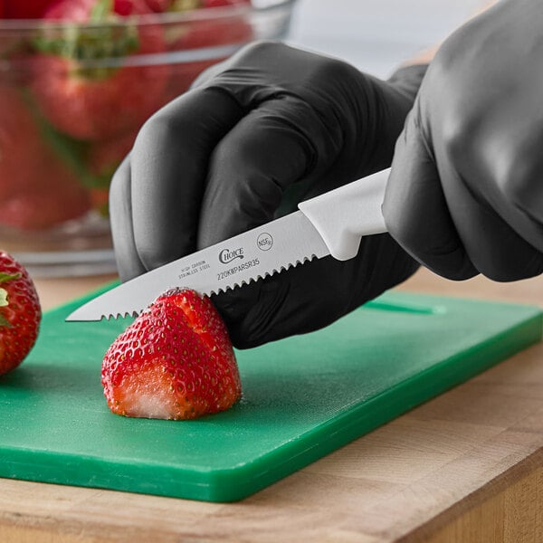 A small serrated edge paring knife with a white handle being used to cut a strawberry on a green cutting board.