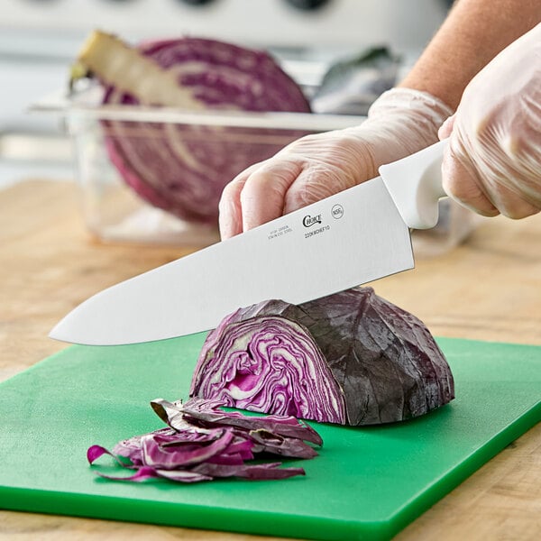 A 10-inch chef knife with a white handle being used to slice a head of purple cabbage on a green cutting board.